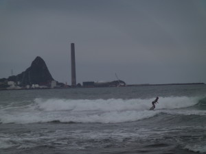 A surfer on Fitzroy Beach. The old New Plymouth Power station sits on the coast (funnel). Paritutu rock is behind - this is the lava plug of an ancient volcano, since eroded away.